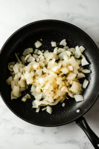 This image shows a skillet over medium heat with olive oil, butter, and diced onions cooking until caramelized. A splash of water may be added to soften the onions faster.