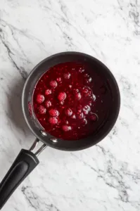 Top-down view of the saucepan over medium heat as the raspberry mixture bubbles gently. The raspberries are releasing their juices, and the sauce is thickening while being stirred, with the texture becoming smoother and thicker.