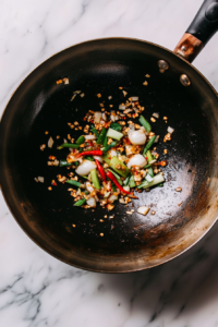 A top-down view of a wok on a white marble cooktop background. Smashed garlic, sliced red chili pepper, and the white parts of scallions are sizzling in oil, releasing their aromatic flavors