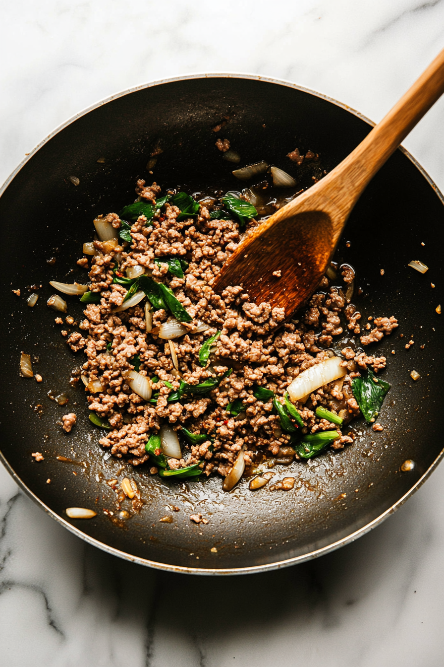 A top-down view of a wok on a white marble cooktop. Ground pork (or chicken) is being stir-fried with garlic, chili, and scallions until fully cooked, turning golden and fragrant.