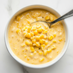 Top-down view of a white bowl filled with creamed corn, served on a white marble cooktop. The creamy sauce coats the corn evenly, and a spoon rests in the bowl, ready for serving. The presentation is clean and simple.