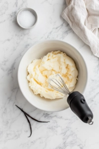 Top-down view of a mixing bowl with butter and ½ cup sugar being creamed using a hand mixer, with vanilla extract and a pinch of salt beside it.