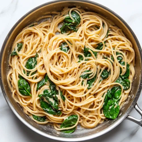 A top-down view of the skillet with cooked whole-wheat spaghetti and wilted spinach being added. Everything is tossed together to coat the pasta evenly with the creamy sauce. The scene shows the vibrant combination of ingredients with the spaghetti and spinach absorbing the rich sauce.