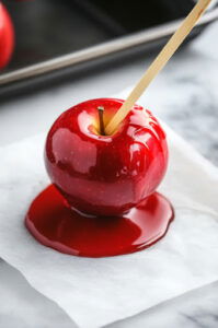 This image shows a person holding an apple by the stick and dipping it into the colored candy syrup, turning it to evenly coat the apple.