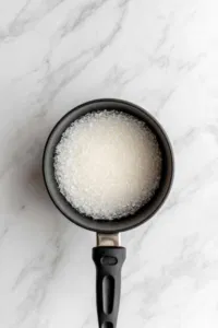 Top-down view of a small saucepan on a white marble cooktop. Water and sugar are being stirred together over medium heat, creating a smooth and clear syrup as the sugar completely dissolves, with the mixture beginning to simmer.