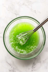 Top-down view of a large mixing bowl on a clean kitchen countertop with a white marble cooktop in the background. Inside the bowl, lime Jello powder is being whisked into boiling water, creating a smooth and vibrant green mixture.