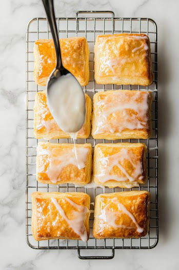 Top-down view of cooled cheese danishes on a wire rack over a white marble cooktop. A smooth glaze made from powdered sugar, vanilla extract, and milk is being drizzled over each danish, focusing on the shiny glaze cascading over the golden pastries.