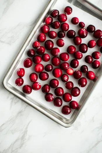 Top-down view of cranberries being removed from the syrup and laid out on a paper towel-lined tray to dry. The glossy cranberries are glistening as they begin to air-dry for 1-2 hours.