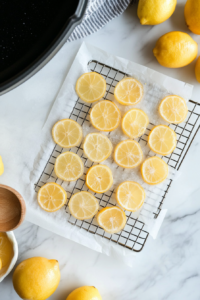 A top-down view of a wire rack lined with parchment paper. Candied lemon slices, translucent and shiny, are laid out in a single layer to dry after being simmered in syrup