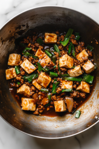 A top-down view of the finished dish in a wok on a white marble cooktop background. The tofu, ground pork (or chicken), and scallions are fully combined, with the sauce coating everything evenly, ready to be served.