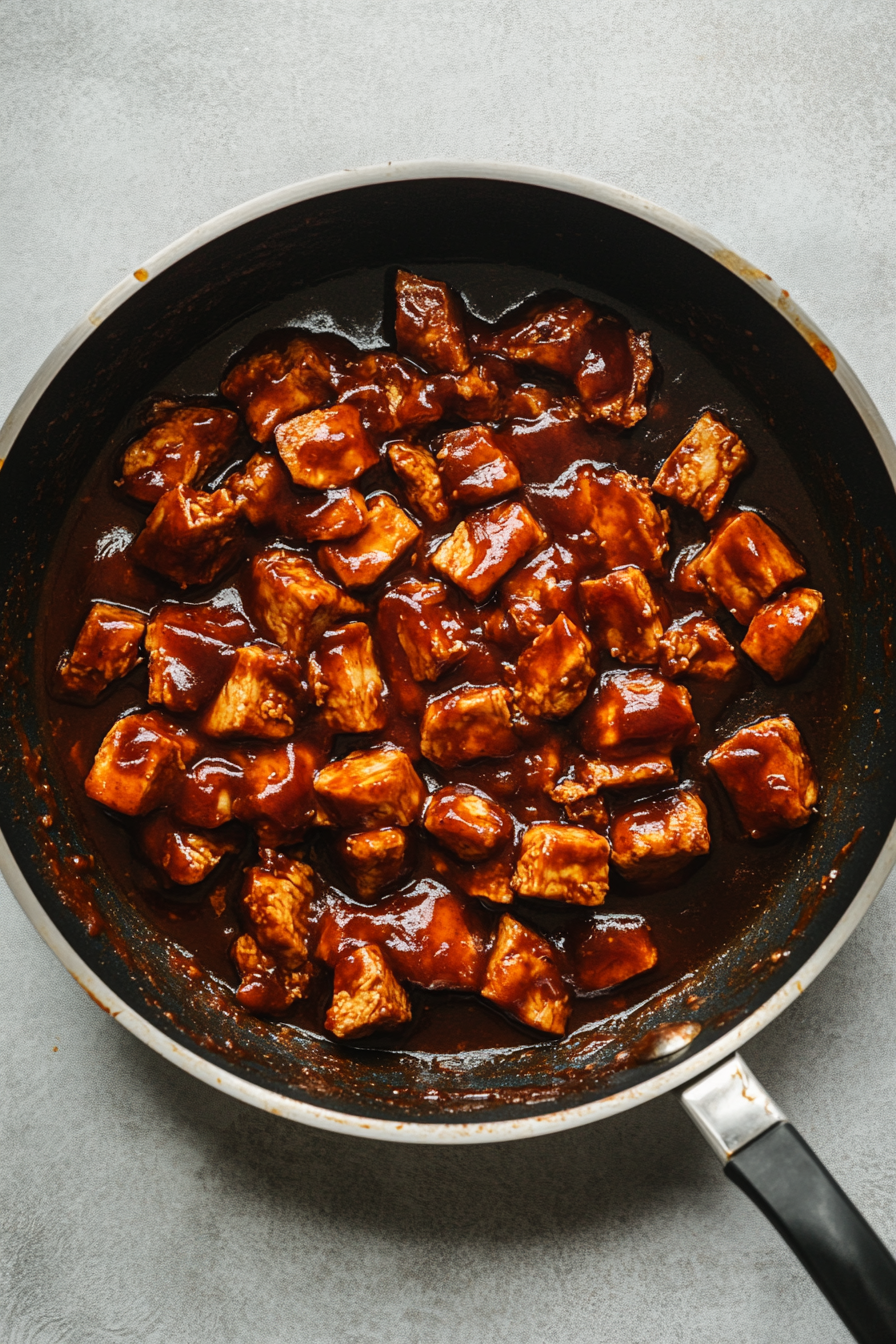 Top-down view of the jackfruit-tofu mixture, glossy and rich with BBQ sauce, after simmering. The final ¼ cup of barbecue sauce is being stirred in, creating a deep, smoky flavor. The scene highlights the thick, flavorful mixture.