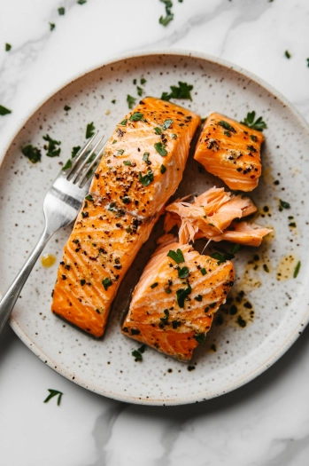 Top-down view of cooked salmon fillets on a plate being flaked into large chunks with a fork. The skin is crispy, and the salmon is perfectly cooked.