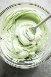 Top-down view of a mixing bowl as Cool Whip is gently folded into the lime Jello mixture. The filling becomes light and creamy, transforming into a pale green color with a smooth and fluffy texture.