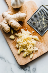 A top-down view of a cutting board on the white marble cooktop. A piece of ginger and garlic cloves are being peeled and finely grated using a small grater. The ginger and garlic are forming a fine paste, with the scene highlighting the fresh, aromatic ingredients being prepared for the sauce.