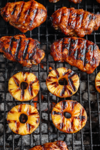 A top-down view of chicken grilling over medium heat, with grill marks beginning to appear on the juicy pieces. Pineapple rings are added to the grill, charring alongside the chicken. The scene captures the caramelizing pineapple and golden chicken on the grill.