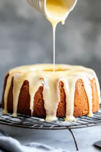 Top-down view of the baked cake on a wire rack, with thick lemon icing being drizzled over the top and dripping down the sides of the cake.