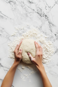 Top-down view of a floured surface with dough being kneaded by hand. The dough is soft and elastic, with flour preventing sticking. The scene captures the kneading process, showing the dough becoming smooth.