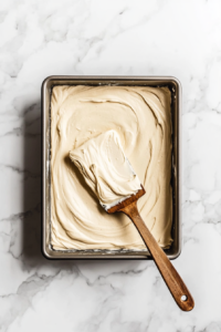 A top-down view of the prepared baking pan with half of the thick batter spread evenly across the bottom using a spatula. The scene focuses on the smooth, even layer of batter.