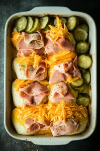 A top-down view of the bottom layer of sweet rolls topped with folded deli ham, shredded aged white cheddar, dill pickles, and Swiss cheese slices, with seasonings of black pepper and garlic powder being added.