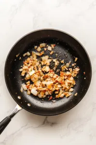 This image shows a large skillet over medium heat with butter and olive oil, minced onions, and garlic being sautéed until softened, seasoned with salt and pepper.