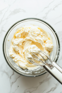 Top-down view of a mixing bowl with cream cheese, icing sugar, and vanilla extract being beaten together with a hand mixer until smooth. The mixture is creamy and lump-free.