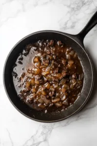 Top-down view of a skillet with caramelized onions, garlic, and white wine reducing. Beef broth, chicken broth, and beef bouillon are being added to the skillet as the sauce simmers and thickens, creating a rich mixture.