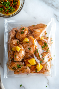 A top-down view of a large ziplock bag on the white marble countertop, with chicken pieces being coated in half of the pineapple marinade. The chicken is evenly covered before sealing the bag for refrigeration, while the remaining marinade is set aside in a small bowl.