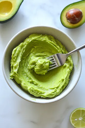 Top-down view of a bowl with a ripe avocado being smoothly mashed using a fork or potato masher.