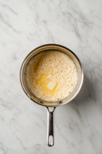 Top-down view of a medium saucepan on a white marble cooktop over medium heat, melting 2 tablespoons of butter with 2 minced garlic cloves sizzling inside. The butter and garlic are gently cooking.