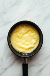 Top-down view of a small saucepan on the white marble cooktop with white chocolate chips, heavy cream, vanilla extract, and honey melting together. Yellow food coloring is being stirred in to create a smooth, creamy yellow mixture, ready to be poured into a lined loaf pan.