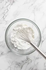 Top-down view of flour, 1 cup sugar, baking powder, and salt being whisked together in a mixing bowl, ready for milk to be added.