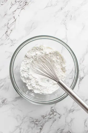 Top-down view of flour, 1 cup sugar, baking powder, and salt being whisked together in a mixing bowl, ready for milk to be added.