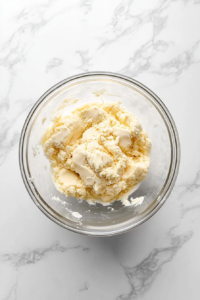 This image shows a mixing bowl filled with butter, sugar, flour, and freshly grated lemon zest being combined to create the lemon cookie dough. A hand is holding an electric mixer to blend the ingredients until a soft, fragrant dough forms, ready to be shaped into cookies.