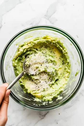 Top-down view of the mashed avocado in a bowl being mixed with parmesan cheese, garlic powder, onion powder, black pepper, and lemon juice using a spoon.