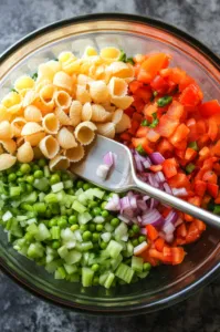 Top-down view of a large mixing bowl containing cooled shell pasta, imitation crab meat, thawed peas, chopped red onion, celery, and red bell pepper, with a spatula ready to stir.