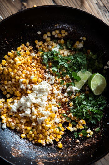 Top-down view of the skillet off the heat, with charred corn, crumbled cotija cheese, mayonnaise, cilantro, lime juice, and chili powder being mixed together. The ingredients are combining into a creamy and flavorful street corn dip.