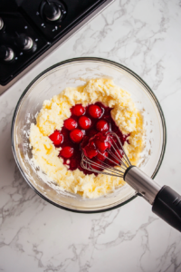 A top-down view of a mixing bowl with lemon cake mix, eggs, and cherry pie filling being combined with a hand mixer. The vibrant mixture is coming together on a clean kitchen countertop with a white marble cooktop background.