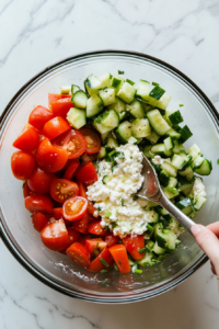 A top-down view of the mixing bowl on the white marble countertop. A spoon is being used to stir the cottage cheese, tomatoes, green onions, cucumbers, and seasoning until well combined. The scene focuses on the vibrant colors and textures of the salad ingredients blending together.