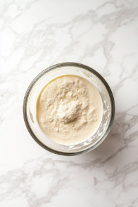 A top-down view of a mixing bowl on a clean kitchen countertop with a white marble cooktop background. In the bowl, unsweetened dairy-free yogurt, nutritional yeast, lemon juice, salt, miso paste, and garlic powder are being whisked together until smooth and creamy.