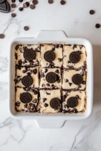 Top-down view of the finished dessert in the baking dish on a white marble countertop. The dessert has cooled and is being cut into squares, showing distinct layers of cookie dough, Oreos, cream cheese, and the crunchy Oreo topping.