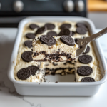 Top-down view of the finished dessert in the baking dish on a white marble countertop. The dessert has cooled and is being cut into squares, showing distinct layers of cookie dough, Oreos, cream cheese, and the crunchy Oreo topping.