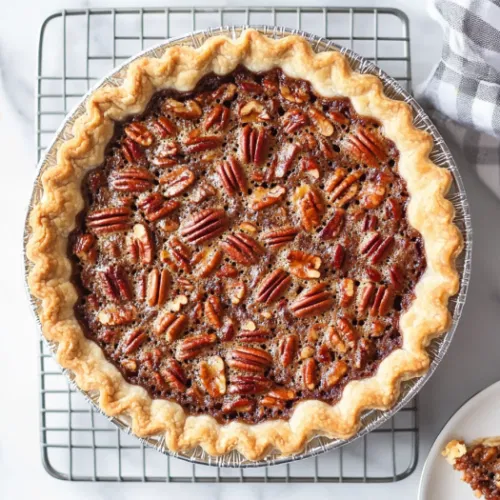 Top-down view of a baked pecan pie cooling on a rack. A golden brown pie with a shiny filling, with a slice cut out and placed on a nearby plate, ready to cool before serving.