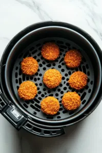 Top-down view of Dorito-coated Babybel cheese rounds being placed in a lined air fryer basket, ready to be cooked at 400°F. The air fryer is sitting on a white marble cooktop.