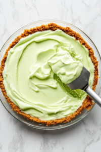 Top-down view of a graham cracker pie crust on the white marble cooktop. The creamy lime Jello and Cool Whip mixture is being poured into the crust, evenly filling it as a spatula smooths the surface.