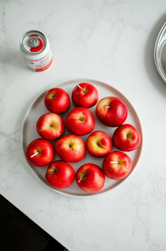 This image shows stemmed apples with craft sticks inserted, ready to be dipped in candy coating. The apples are placed on a lightly greased cookie sheet.