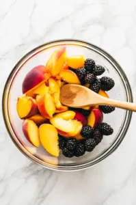 Top-down view of peeled peaches, blackberries, lemon juice, cinnamon, and ¼ cup sugar being stirred together with a wooden spoon in a bowl.