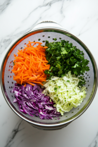 Top-down view of a large colander with grated carrot, thinly sliced green cabbage, red cabbage, red onion, and chopped parsley, sprinkled with kosher salt, set over a bowl on a clean kitchen countertop.