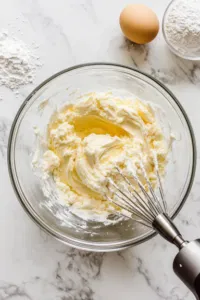 Top-down view of softened butter, golden caster sugar, eggs, Greek yogurt, vanilla extract, and lemon zest being mixed with an electric mixer in a large bowl. Flour, baking powder, and salt are set aside, ready to be folded in.