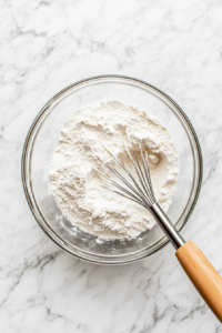 A top-down view of a medium mixing bowl with all-purpose flour, baking soda, banana cream instant pudding mix, and kosher salt being whisked together. The scene shows the dry ingredients being evenly combined.