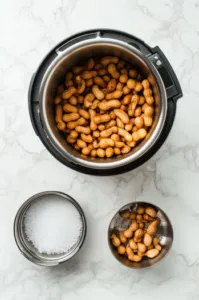 Top-down view of raw peanuts in their shells, salt, and liquid smoke measured out and placed beside an Instant Pot on a white marble countertop.